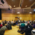 A wide shot of attendees sat in the audience at a conference watching a speaker at a podium